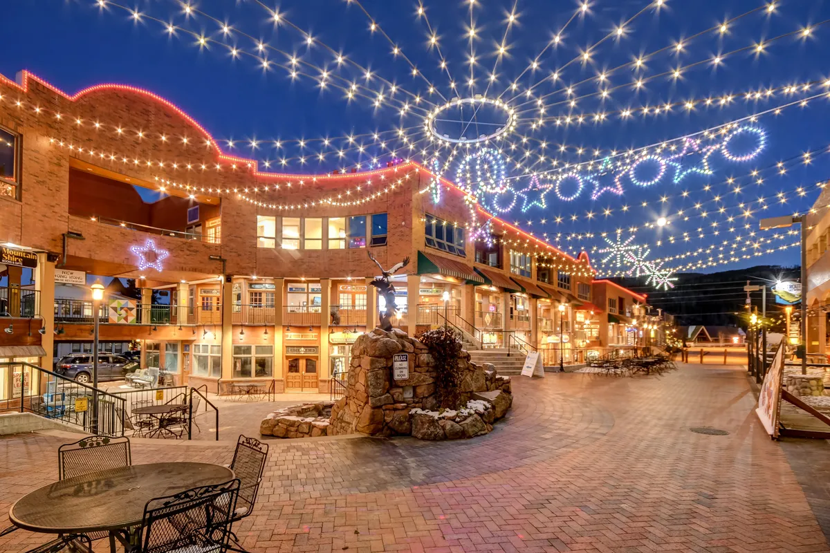 A vibrant exterior image capturing a lively local plaza adorned with string lights and decorative stars, with people strolling, dining, and shopping amid a festive atmosphere around a central fountain.