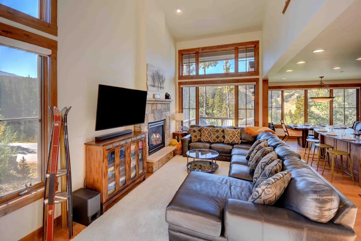 This image captures the open-concept living area with a large sectional sofa, a glowing fireplace, and floor-to-ceiling windows that flood the space with natural light. The dining area is visible in the background, and the overall atmosphere is cozy and inviting, with mountain-inspired décor touches.