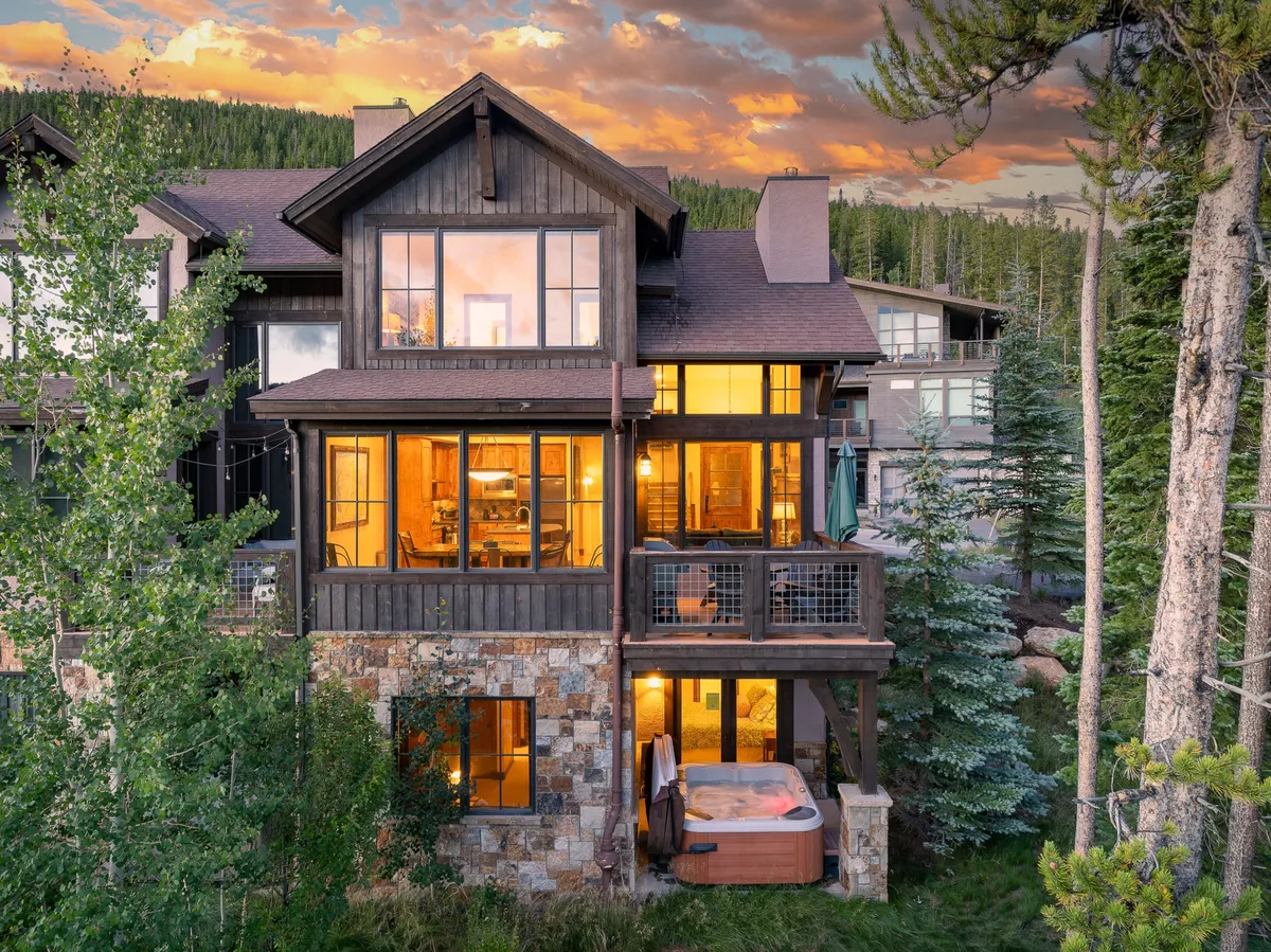 The image showcases the stunning exterior of a luxury single-family villa nestled among towering trees in Winter Park, Colorado. The home features a beautiful stone facade, expansive windows aglow with warm light, and a spacious deck with a private hot tub. The inviting outdoor space is perfect for gathering with friends or relaxing in nature, while panoramic mountain views and access to nearby trails set the stage for adventure and relaxation.