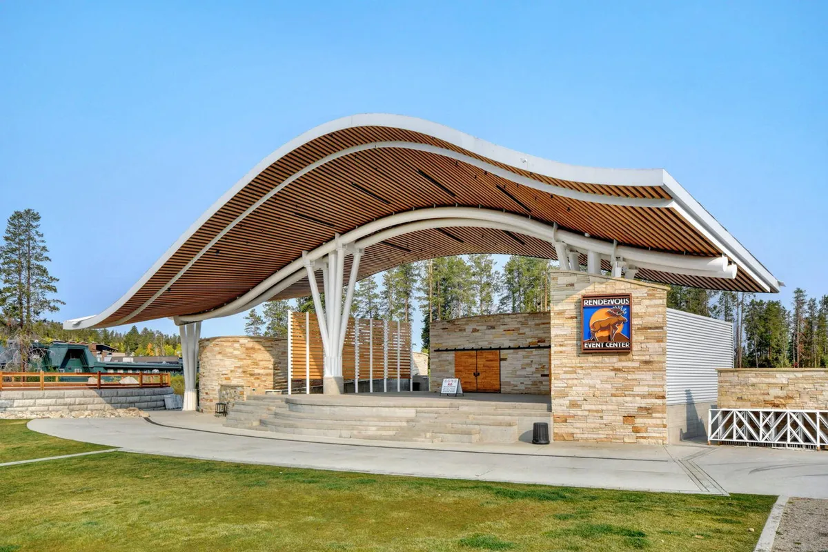 A dynamic exterior view of a unique community venue with a wave-like roof, stone facade, and large grassy area, suggesting a hub for concerts, outdoor festivals, and family gatherings.
