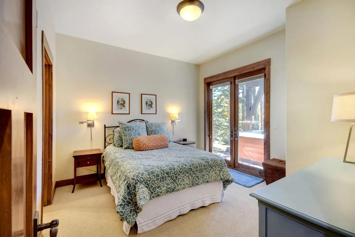 A serene bedroom featuring a king-sized bed adorned with patterned bedding, two bedside lamps, and framed artwork. Double doors lead to an outdoor space, allowing natural light to fill the room and offering a tranquil view.