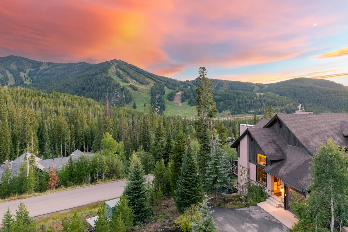 An exterior shot emphasizing the modern architecture of the property set against a backdrop of a glowing sunset, forest greenery, and majestic mountains. The scene suggests the property's prime location for enjoying nature and breathtaking views.