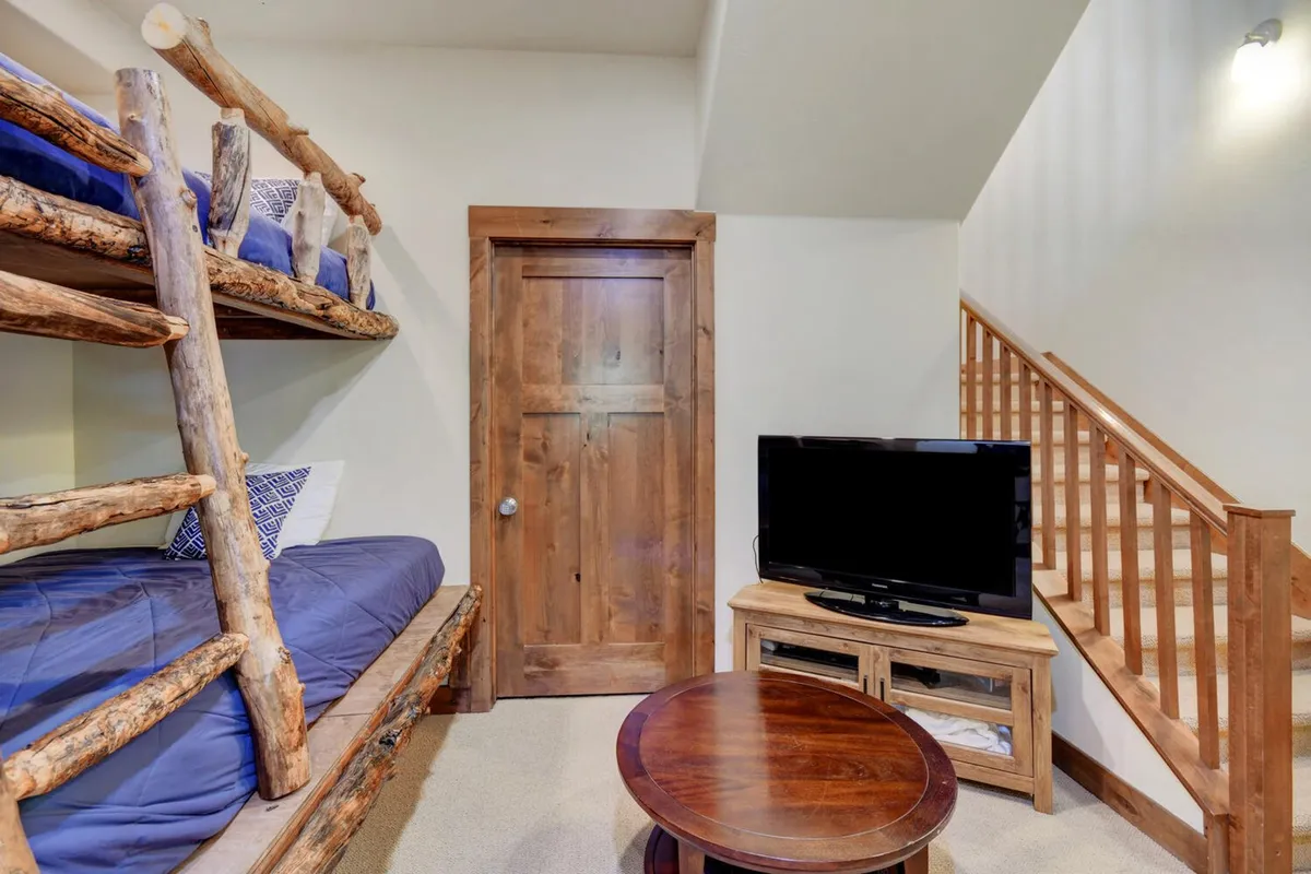 A cozy bunk room equipped with custom bunk beds, a television, a wooden door, and stairs. The space is inviting and playful, perfect for children or groups to unwind with games or TV after a day of adventure.