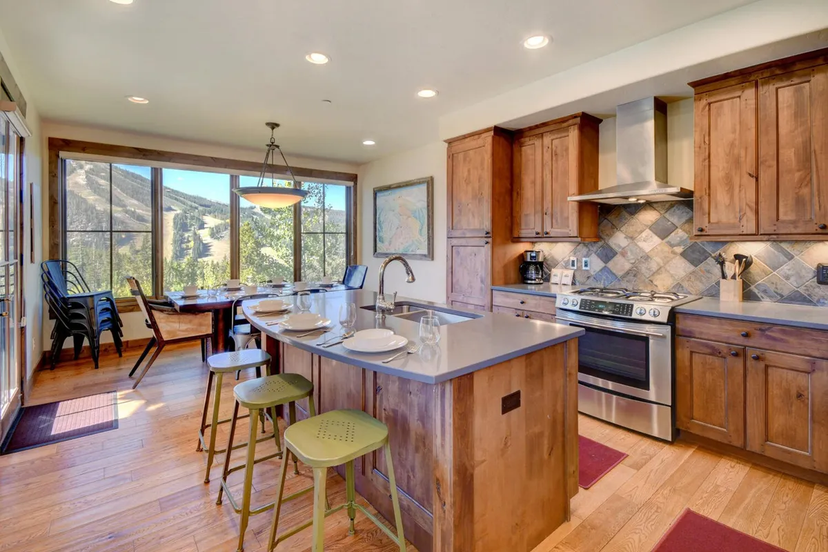 A view of the kitchen island, dining table, and expansive windows, this image highlights how the kitchen seamlessly merges with the dining and living areas. It features natural light pouring in and modern amenities, illustrating the open concept and social flow described.